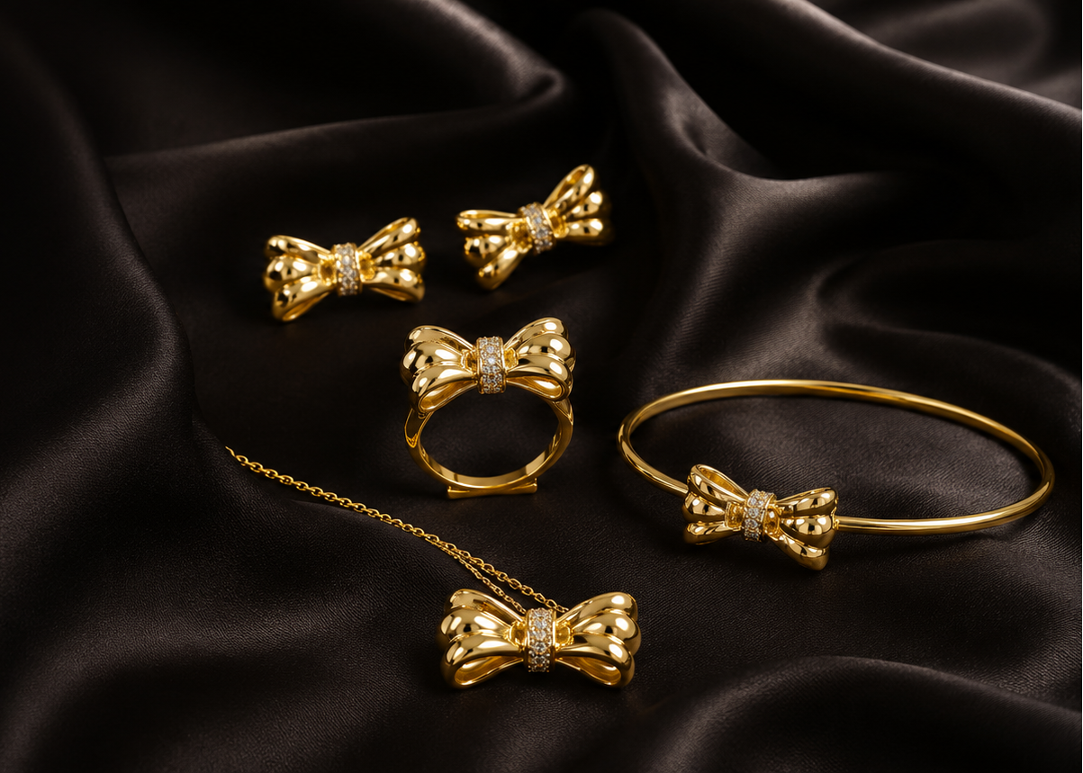 A set of gold jewelry featuring multiple bow-shaped pieces, including earrings, a ring, a bracelet, and a necklace, arranged on a draped black satin fabric background.